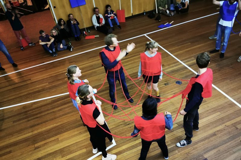 6 players in red jerseys are complexly connected by red cords. They are standing on a wooden floor surrounded by an audience with blue and red picket signs.