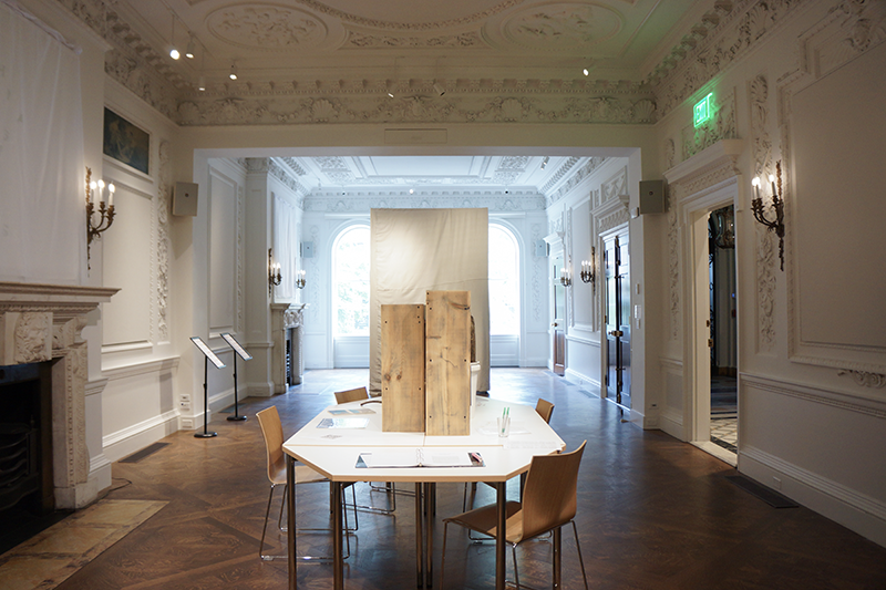 An art installation in the Salon at Goethe Institut, Boston. A high ceilinged white room filled with decorative wall plasters. A dirty library on raw wood shelves on a white octagonal table. Plasters translated into emojis, shrouded mirrors and a dirt shrine in a ten by six by six canvas cube.