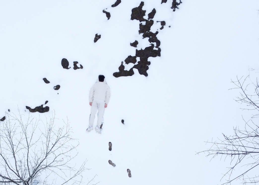 An aerial view of a body, face down, dressed in white, in a snowy landscape, the branches of two trees reach into the bottom corners of the frame and the dark, melted spots reach down from the top to the center of the frame
