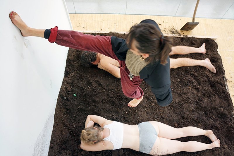 two nearly naked bodies lie facedown in a square of dirt, while a third person extends a dirty foot onto the white wall on one side of the dirt bath.