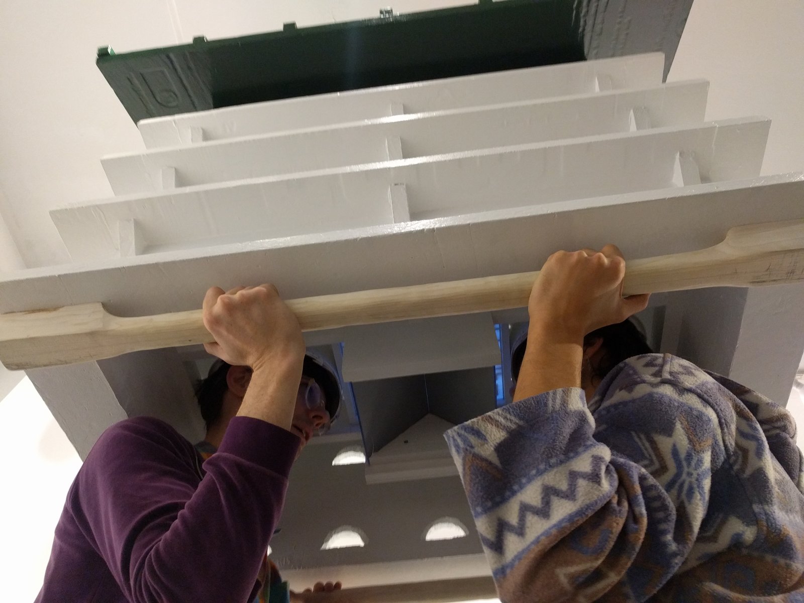 2 people stand facing each other inside a giant white & green bird house. Between them a pair of mirrors reflect a maze.