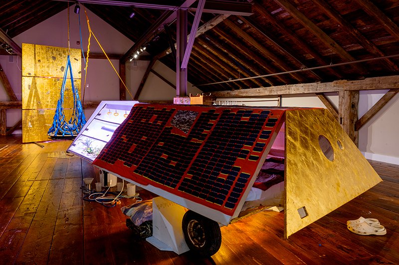 In the foreground a satellite turned into habitation module with greenhouse sits on a pair of wheels, its front half lifted off the ground by a cargo net full of pillows, behind the net a golden climbing wall covered in body parts