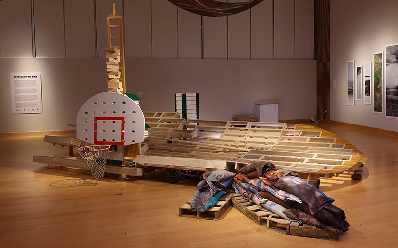 Poplar rectangles come together to make a boat shaped like the key or free throw lane of a basketball court. The semicircle front of the boat shape extends into a sliver of stained oak court flooring. From beneath the oak flooring skinny pallets extend, mimicking the branching of the Shenandoah River. The pallets hold 23 sand bags tucked into images from just above and below the Shenandoah River at Port Republic where the gundalows were built and launched. To the left of the sandbags a displaced segment of a swift tower, turned on its side, stands on four legs and holds a tall skinny cart, stacked with wooden forms for casting clay letters. A basketball hoop is bolted to the side of the tower and a fan shaped backboard rests on top of the hoop and leans against the cart. The backboard is pierced with a grid of starling resistant entry holes, that are used on purple martin houses to avoid starlings entering and destroying the martin’s nests. Three of the holes have doll house size models of the 72 room purple martin hotel built for the first Dirtball court. These doll house models are the size of a single room in the original purple martin hotel. Inside these models are images of Dirtball courts and the architectural remnants of the repurposed Shenandoah River gundalows. Another section of a chimney swift tower rises behind the stern of the boat, against a far wall a white chest freezer.