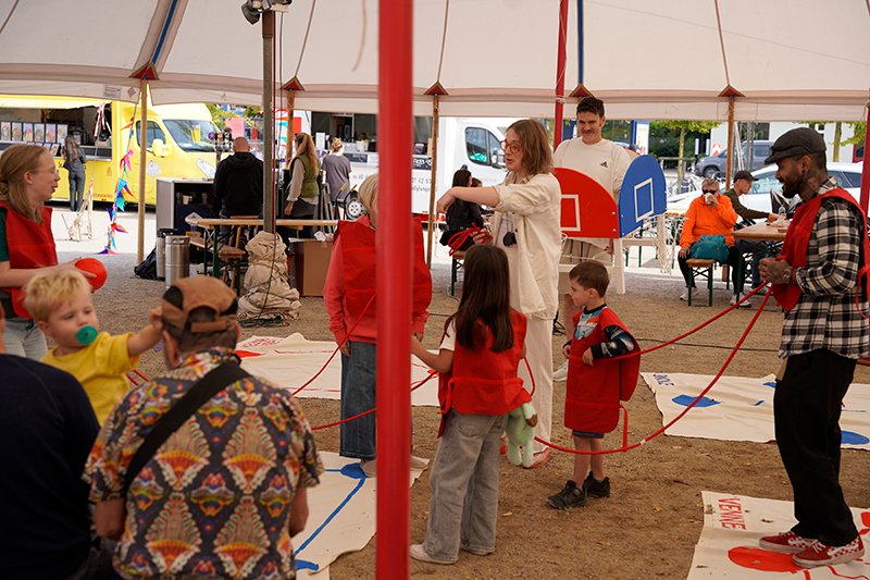 Inside a circus tent, players in red jerseys are connected by red ropes. Painted Diagrams are on the floor, a person wears a multi-goal basketball hoop.