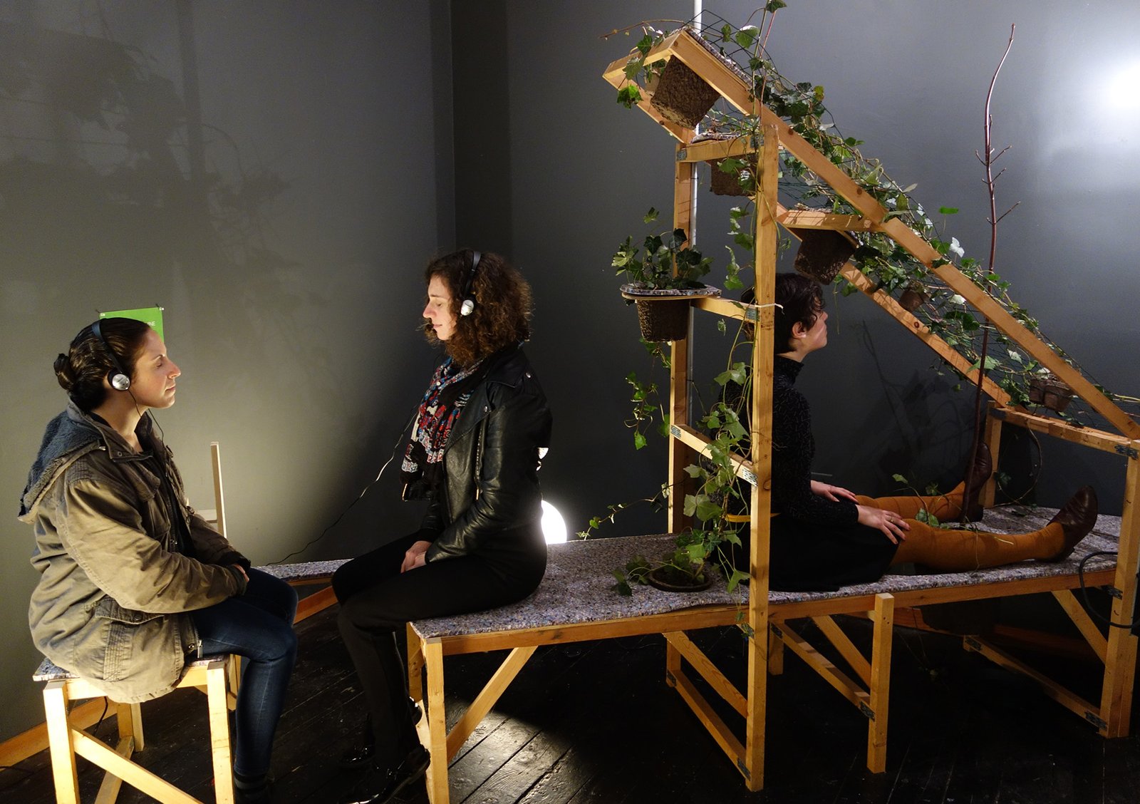 A piece of custom furniture made of wood and felt holds a host of plants and 3 humans. 2 of the people are facing each other and listening to a meditation on headphones. The third is surrounded by ivy plants and a young tree.