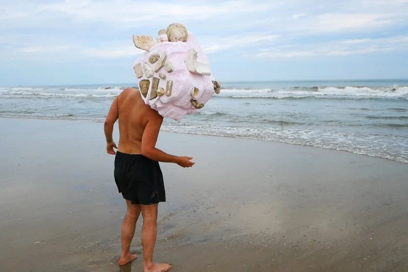 A person in a black bathing suit stands on the beach in front of the waves. Their head is replaced by giant pink foam and discarded river styrofoam helmet with a maze inside.